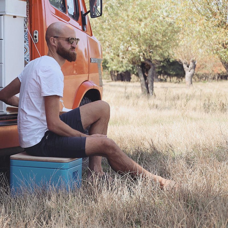 A bearded man sitting on a Trelino portable composting toilet next to an orange campervan.
