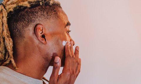 Man with locs applying facial moisturizer in bathroom mirror