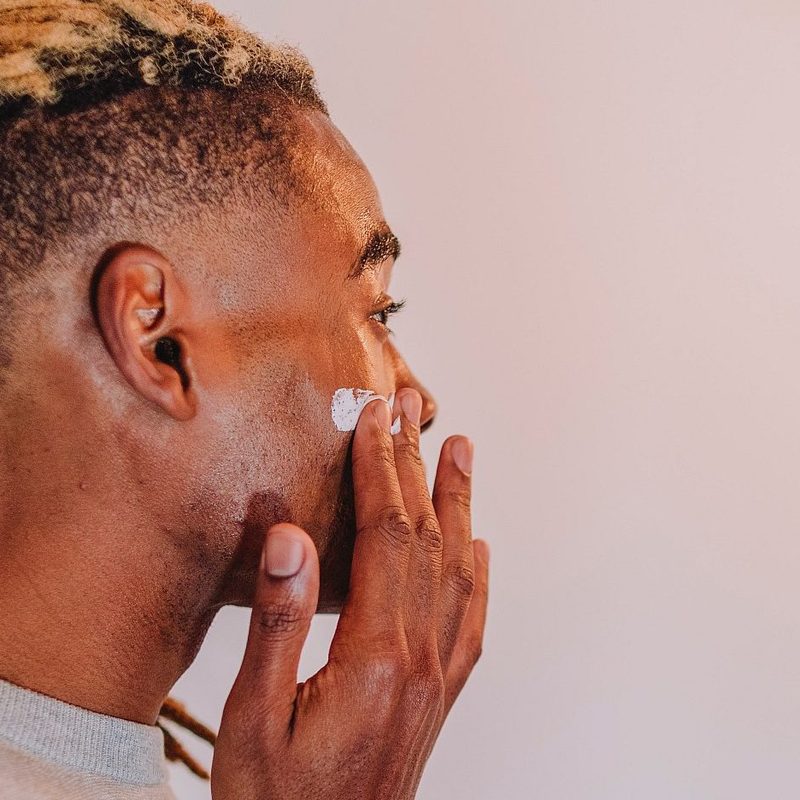 Man with locs applying facial moisturizer in bathroom mirror
