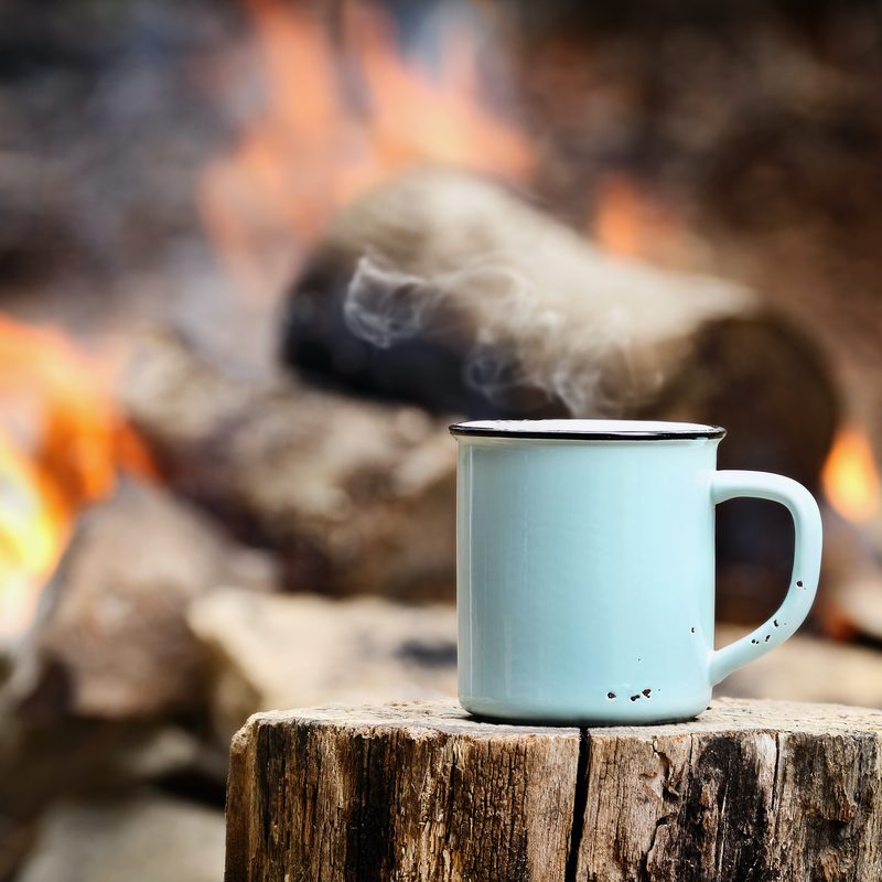 Blue enamel cup of hot steaming coffee sitting on an old log by an outdoor campfire. Extreme shallow depth of field with selective focus on mug.