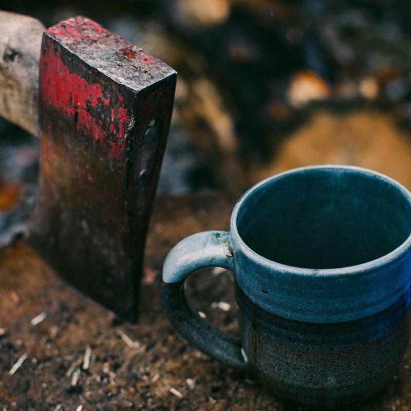 Red-headed ax buried into a tree stump next to a blue coffee mug in the outdoors