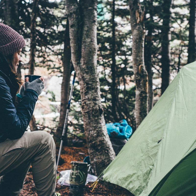 Young woman enjoying a cup of coffee, while sitting near a tent in the outdoors.