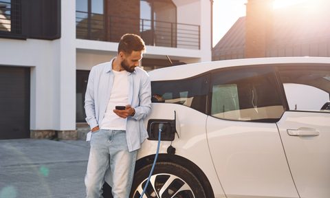 Charging the vehicle near the house. Holding smartphone. Young stylish man is with electric car at daytime.