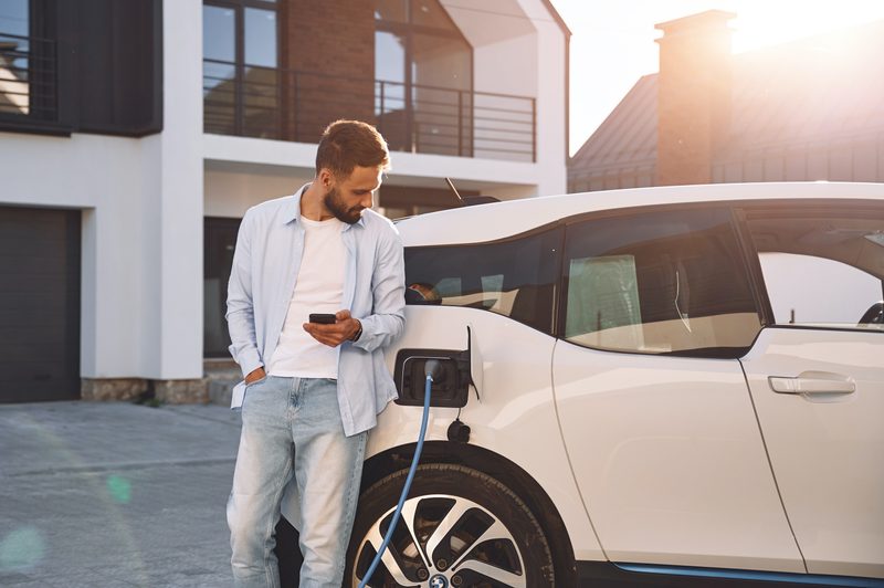 Charging the vehicle near the house. Holding smartphone. Young stylish man is with electric car at daytime.