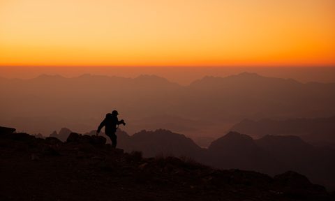 Silhouette of a hiker standing on top of a mountain with a bright orange sky in the background