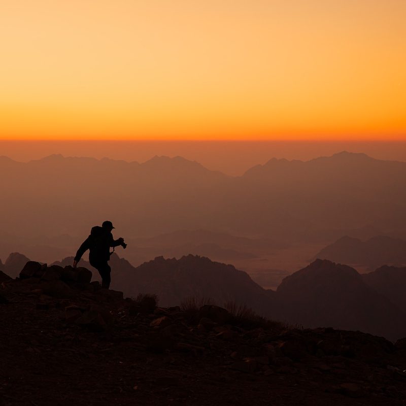 Silhouette of a hiker standing on top of a mountain with a bright orange sky in the background.