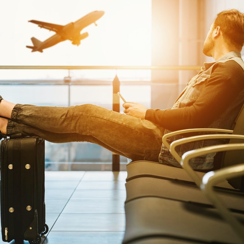 Seated man with feet propped up on luggage looks longingly out airport window at plane taking off