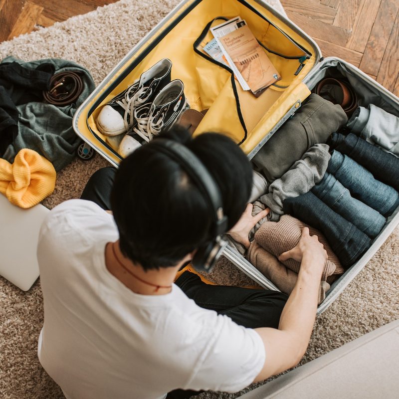 Top view of a man wearing headphones while packing a suitcase with clothing