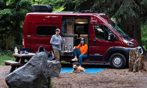 Couple standing outside near the open door of a Winnebago Solis Pocket 36B RV parked at a campsite.