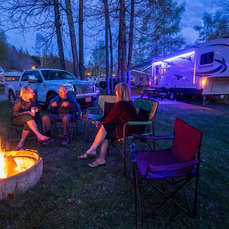 Three people sit around a fire with a lit up RV in the background.