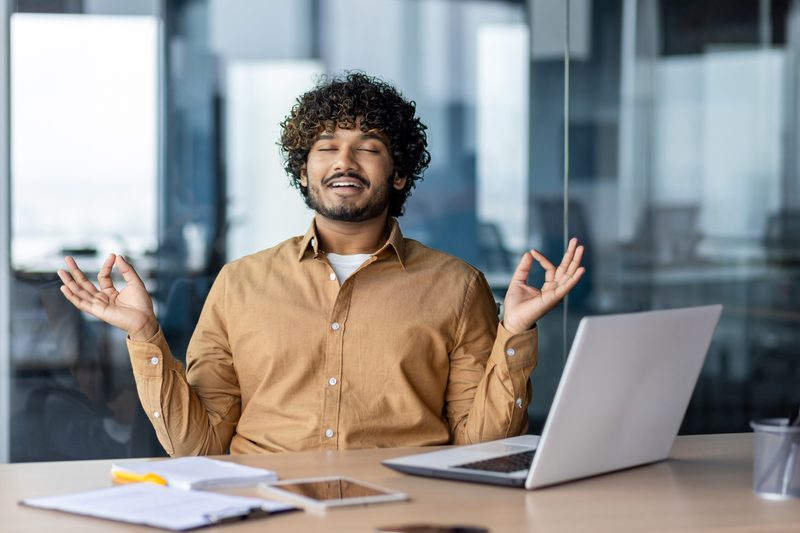 Man resting in office relaxing, businessman meditating in lotus position at workplace, with laptop doing yoga during break.