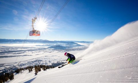 Skier heading downhill at Jackson Hole Mountain Resort, with the sun and aerial tram in the background.