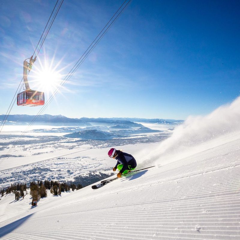 Skier heading downhill at Jackson Hole Mountain Resort, with the sun and aerial tram in the background.