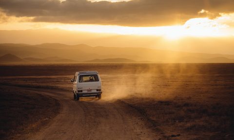 White campervan driving a dusty road in a hot desert landscape.