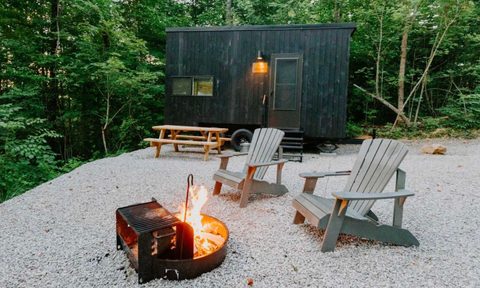 A small, black wood-paneled tiny house in the woods with a firepit and chairs in the foreground