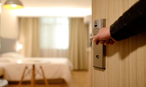 A hand opening a hotel room door with the room interior in the background