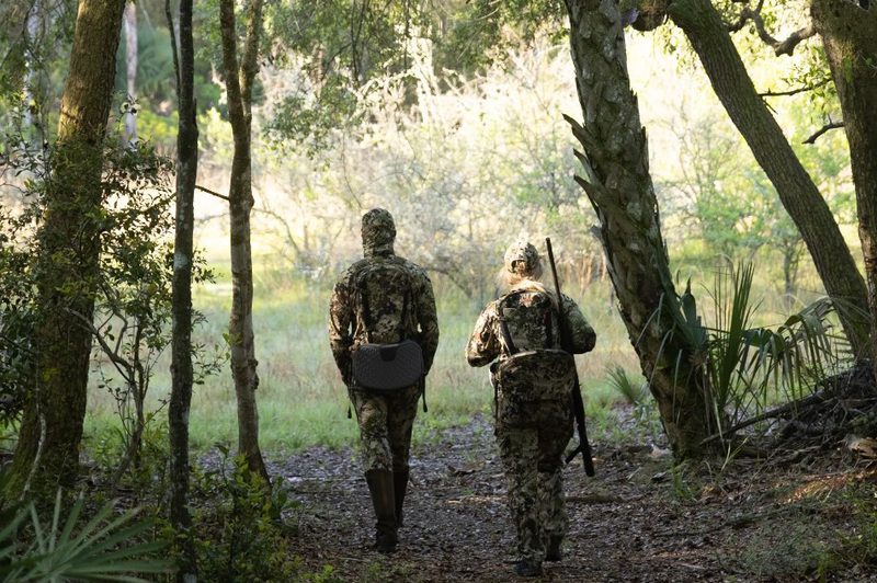 Two hunters walking through a swampy wilderness in camouflaged, insect-repellent clothing.