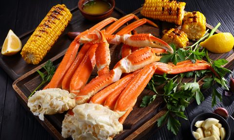 Snow crab legs served with melted butter, garlic cloves, lemon slices, grilled corn in cobs and fresh parsley on a wooden cutting board