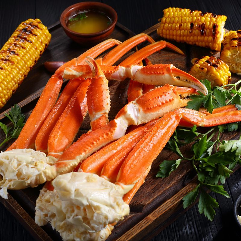 Snow crab legs served with melted butter, garlic cloves, lemon slices, grilled corn in cobs and fresh parsley on a wooden cutting board