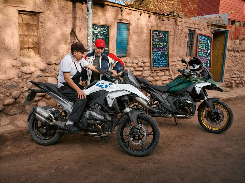 Two 2024 BMW R 1300 GS adventure touring motorcycles parked outside a cafe with a cook sitting on one as the owner looks on.