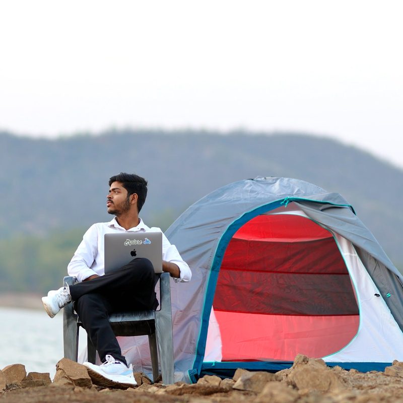 A man sitting while holding a laptop at his campsite off a body of water on a cloudy day