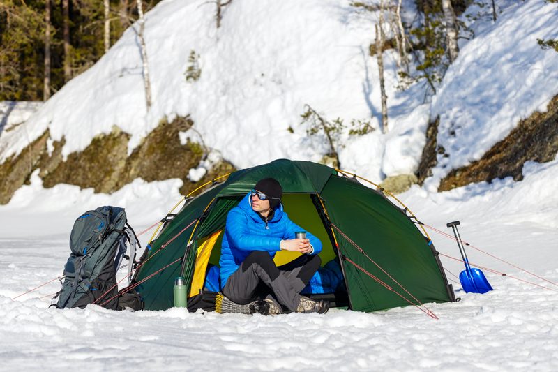 Happy man drinking tea in a green tent in the forest. Nearby are a backpack and a snow shovel. Beautiful sunny spring day.