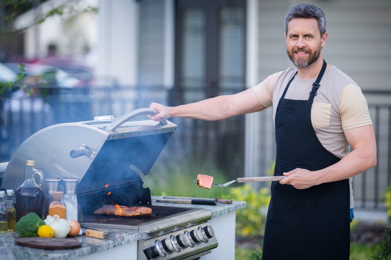 Male chef grilling and barbequing in garden. Barbecue outdoor garden party. Handsome man preparing barbecue meat. Concept of eating and cooking outdoor during summer time