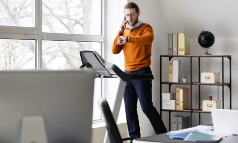 Young man talking by mobile phone while running on treadmill in office