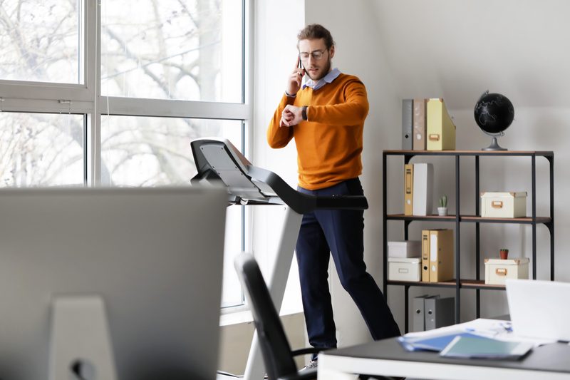 Young man talking by mobile phone while running on treadmill in office