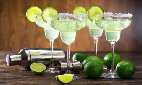 Four glasses of freshly made Margarita cocktail decorated with limes on wooden table with shaker and limes.