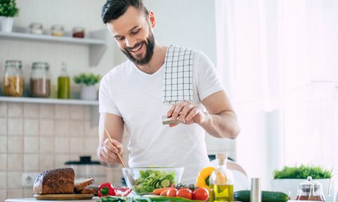 Handsome happy bearded man is preparing wonderful fresh vegan salad in the kitchen at home