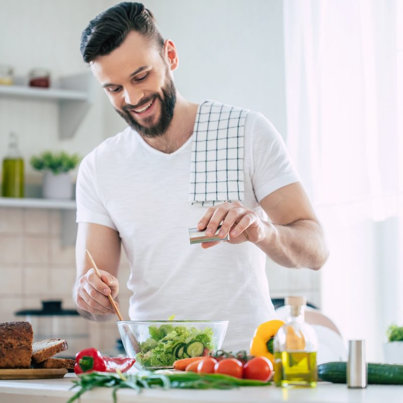 Handsome happy bearded man is preparing wonderful fresh vegan salad in the kitchen at home