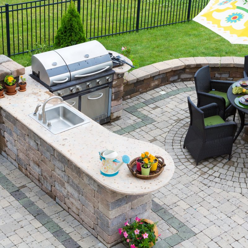 High angle view of a stylish outdoor kitchen, gas barbecue and dining table set for entertaining guests with formal place settings and flowers on a paved patio