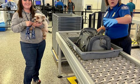 A woman holding her dog while the pet carrier does through the X-ray.