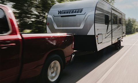 Red pickup truck towing Winnebago Access travel trailer down the road.