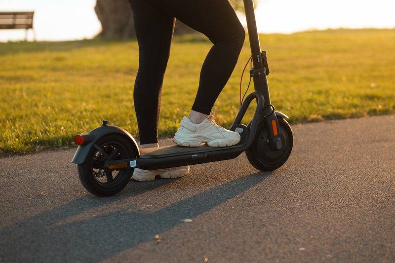 A person standing on a NAVEE V40 Pro electric commuter scooter with their right foot on the deck and left foot on a paved path.