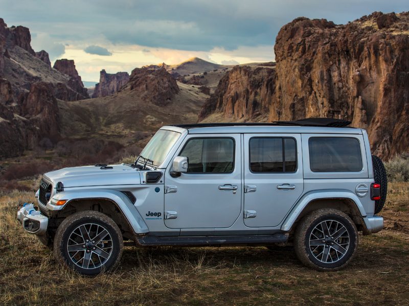 Silver 2024 Jeep Wrangler High Altitude 4xe parked on wild grass on a rise with hills in the background.