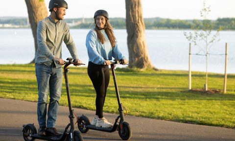 Two people wearing helmets and riding NAVEE V40 Pro electric commuter scooters on a paved path through a grassy area beside a lake.