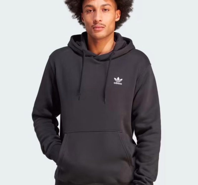 A model wears a black Adidas Trefoil Essentials Hoodie against a gray background.