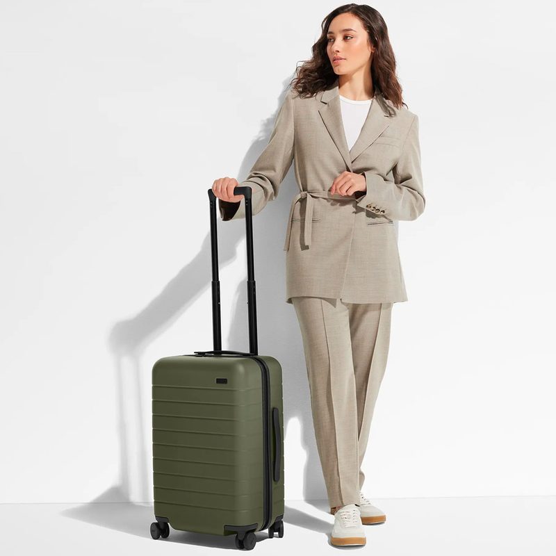 A woman stands next to the Away Travel Carry-On suitcase in olive green