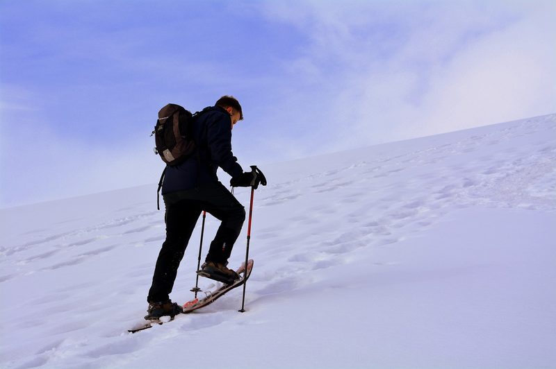 Male hiker walking up a snowy hill in snow shoes and trekking poles