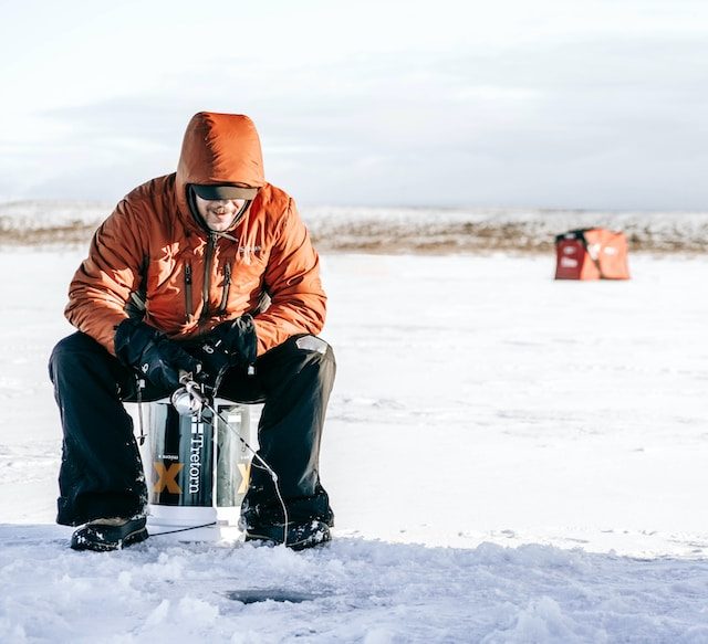 A man in an orange jacket using an auger and ice fishing