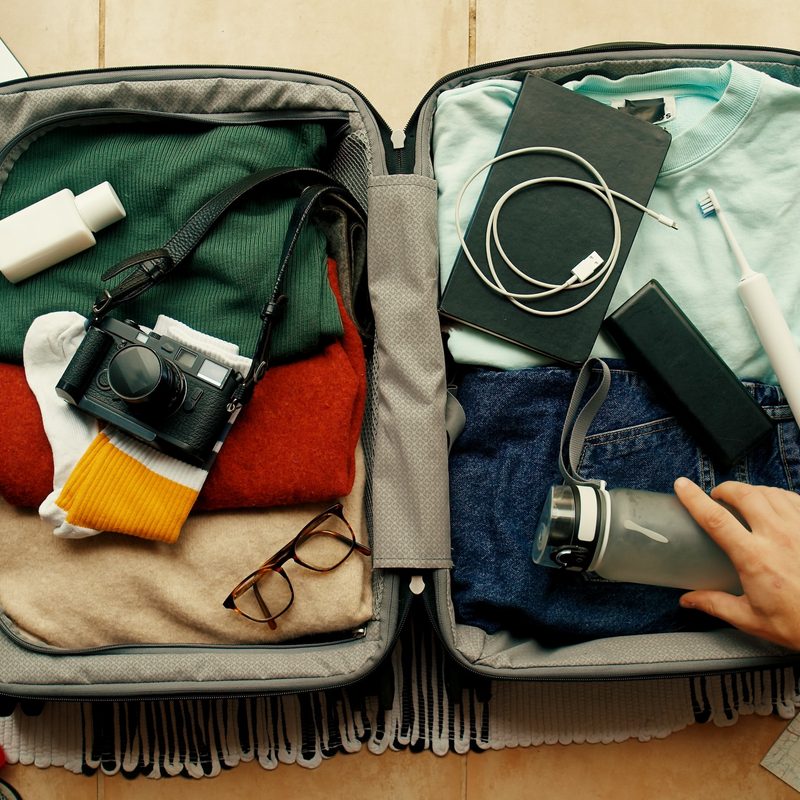 Man with tatted arm packs suitcase for adventure travel trip. Packing bag with camera, laptop, portable battery charger and electric toothbrush.