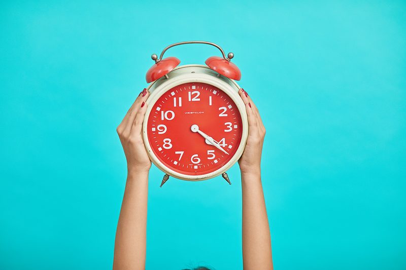 Hands holding red and white alarm clock up in the air with blue background