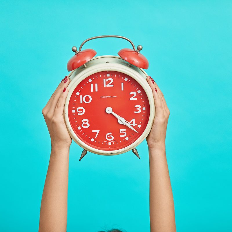 Hands holding red and white alarm clock up in the air with blue background