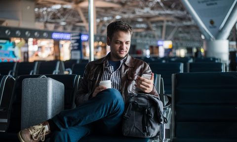 Portrait of handsome smiling man in casual wear holding his luggage and messaging through his mobile phone while sitting in the hall of the airport.