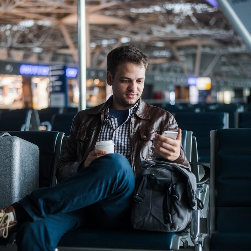 Portrait of handsome smiling man in casual wear holding his luggage and messaging through his mobile phone while sitting in the hall of the airport.