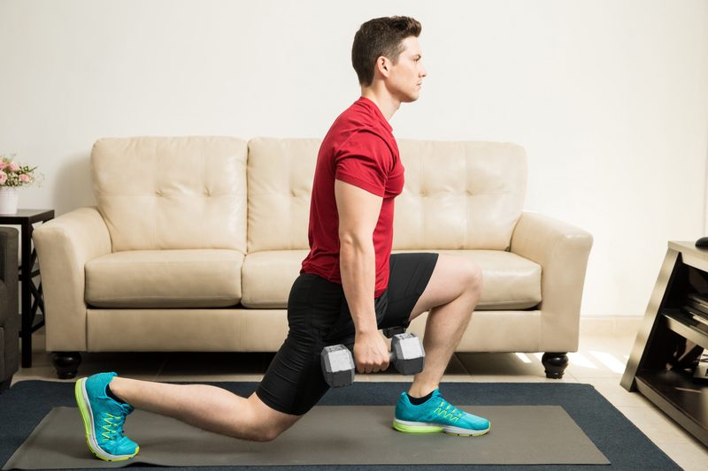 Profile view of a young and fit man doing kneeling lunges with a pair of dumbbells at home
