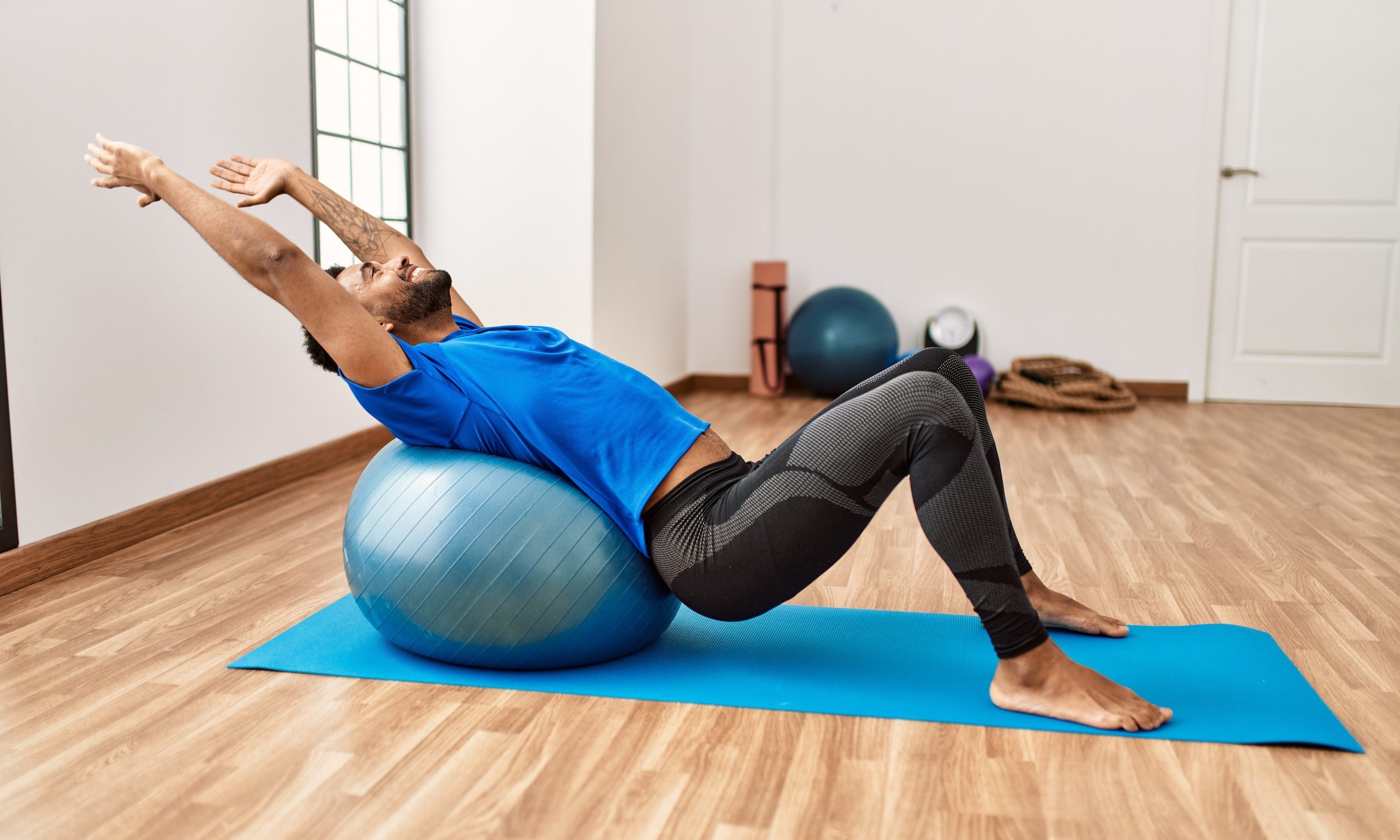 Handsome hispanic man doing exercise and stretching on yoga mat, practicing flexibility with pilates ball at the gym