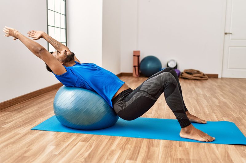 Handsome hispanic man doing exercise and stretching on yoga mat, practicing flexibility with pilates ball at the gym